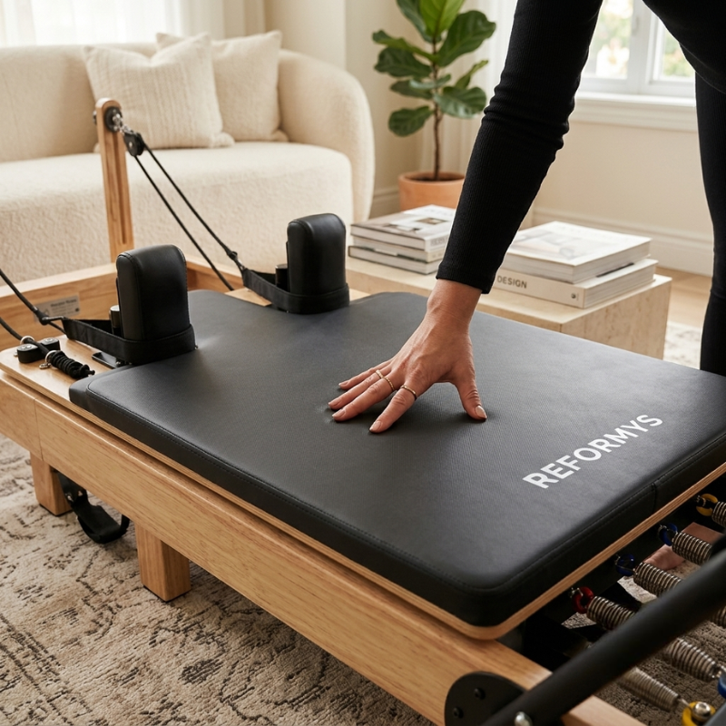 A person's hand resting on the Reformys Pilates Reformer Mat in Black that's sitting on a Pilates reformer machine inside a home lounge